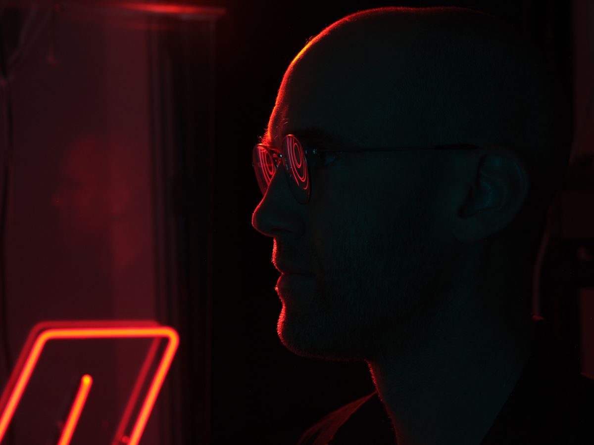 A silhouetted profile of a person wearing glasses illuminated by dramatic red and orange neon lighting in a dark urban setting. The atmospheric lighting creates a moody, cyberpunk-style scene with glowing neon signs visible in the background.