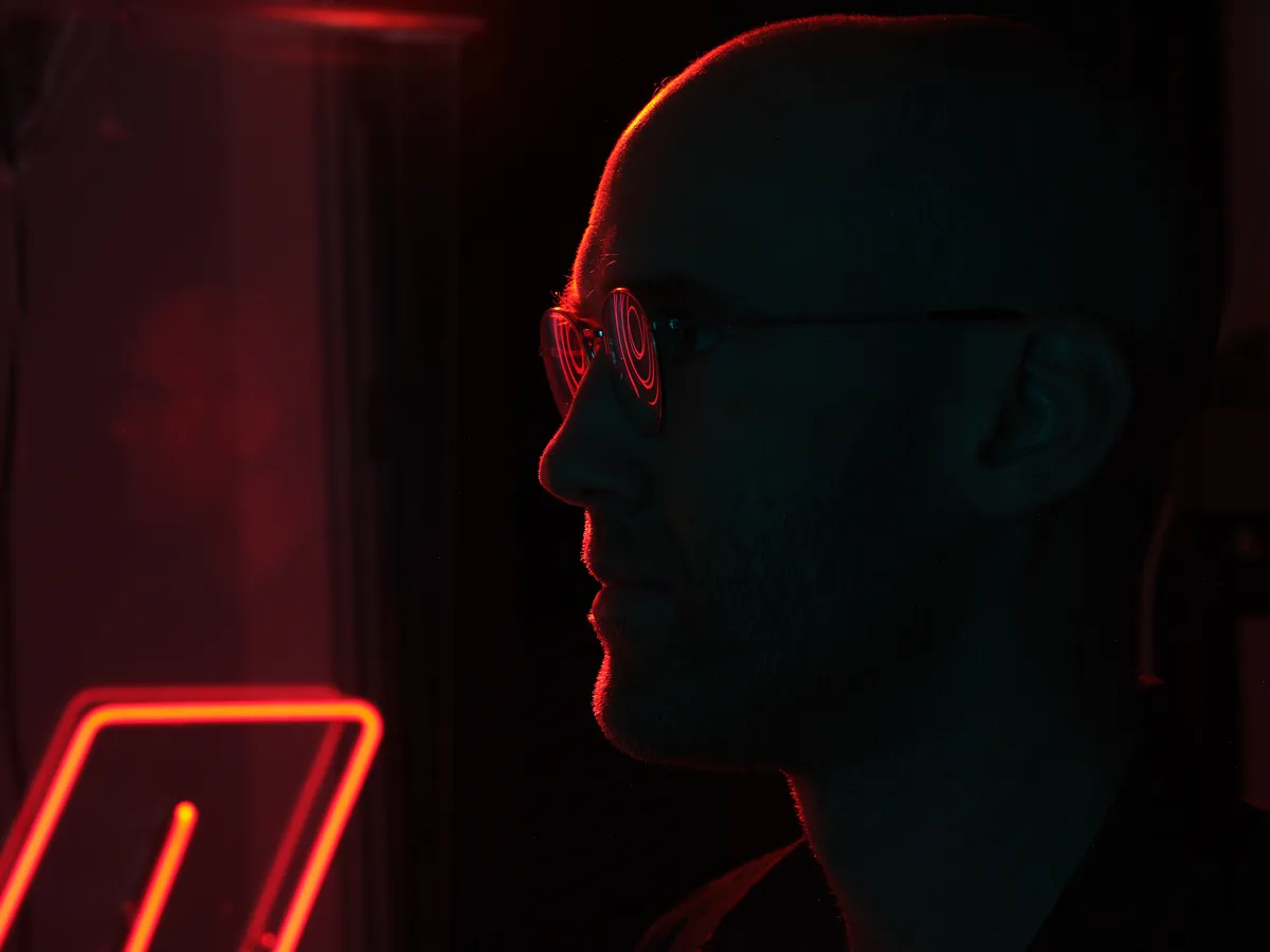 A silhouetted profile of a person wearing glasses illuminated by dramatic red and orange neon lighting in a dark urban setting. The atmospheric lighting creates a moody, cyberpunk-style scene with glowing neon signs visible in the background.