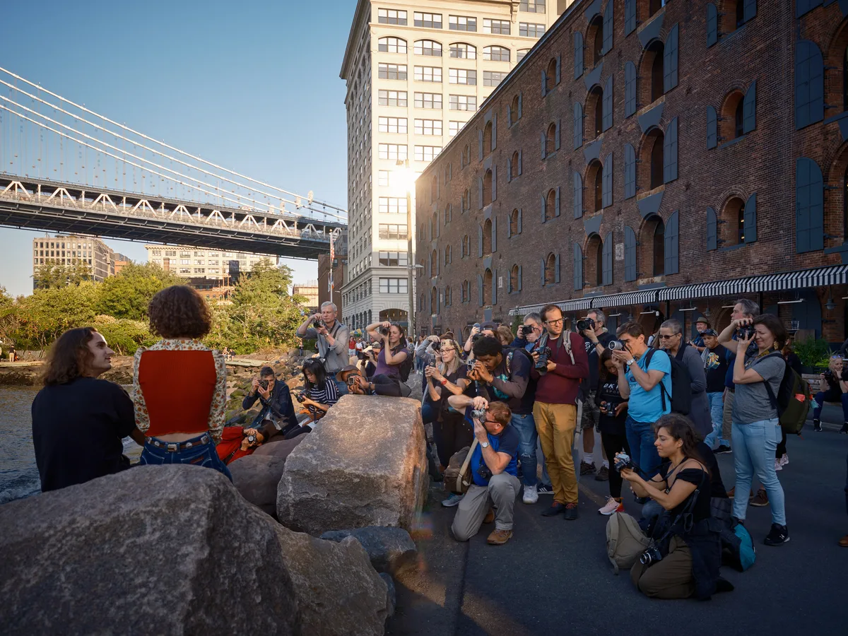 A vibrant gathering of photographers and onlookers assembles along the cobblestone waterfront beneath the Manhattan Bridge in Brooklyn's DUMBO neighborhood. The late afternoon light bathes the historic brick warehouses and modern high-rises in warm golden tones, while dozens of camera-wielding enthusiasts focus their lenses on subjects positioned among weathered granite boulders. The iconic suspension bridge cables stretch dramatically overhead, framing this quintessential New York photography scene where industrial heritage meets contemporary urban culture.