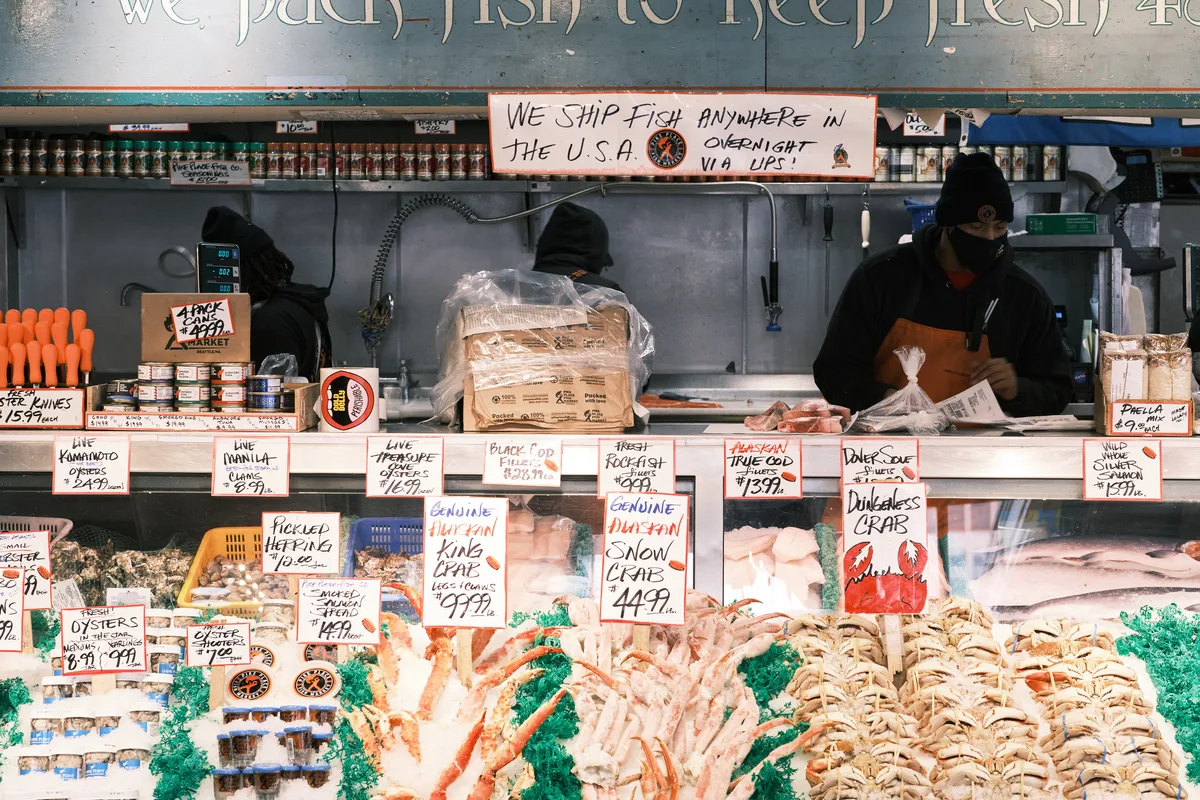 Behind the iconic seafood counter at Seattle's Pike Place Fish Market, workers in black hoodies tend to their daily catch beneath handwritten price signs and shipping notices. The bustling display showcases an abundance of fresh seafood arranged on crushed ice, from Dungeness crab to Alaskan true cod, while bright orange price tags punctuate the scene. The industrial stainless steel workspace and vintage signage create an authentic portrait of this legendary Seattle marketplace, where maritime tradition meets urban commerce.