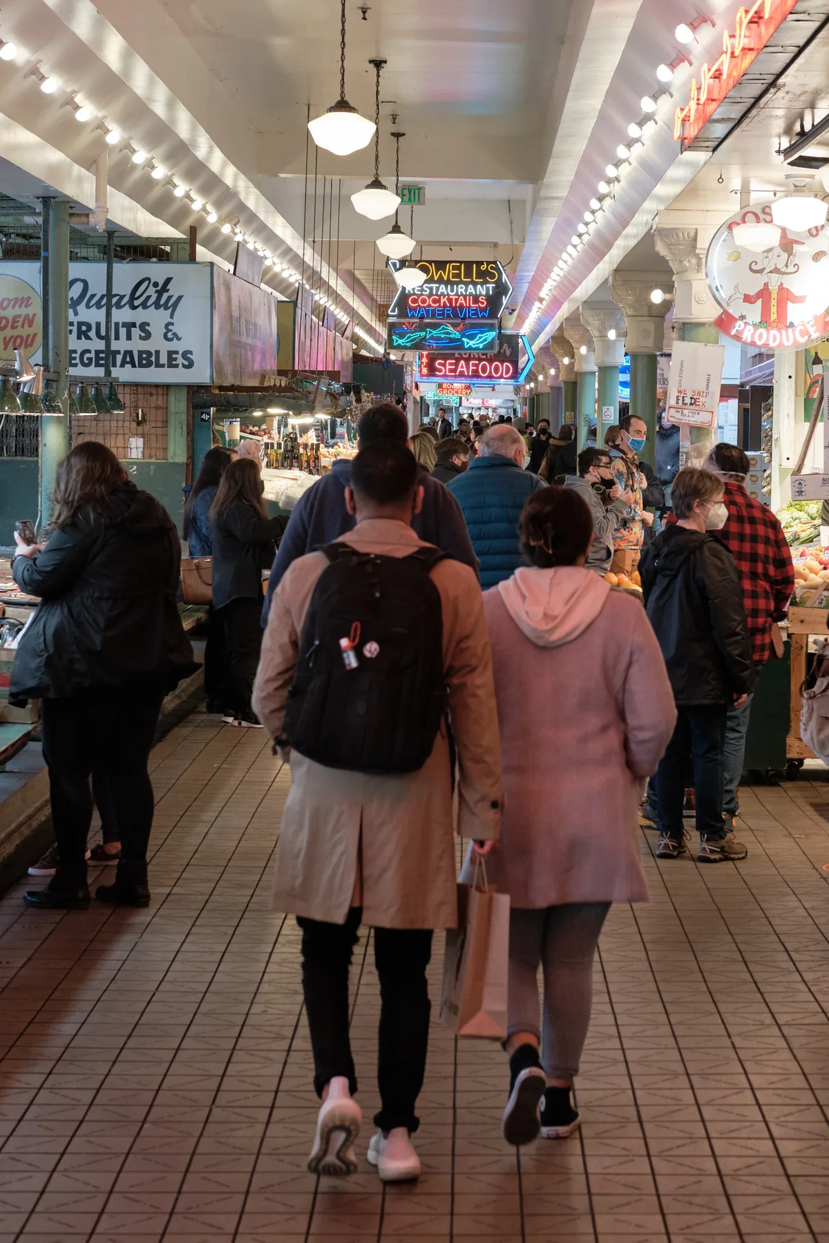 Crowds of visitors navigate the iconic main corridor of Pike Place Market, Seattle's historic public market. The scene captures the market's characteristic energy as shoppers meander past vintage neon signs, including Lowell's Restaurant and quality produce stalls. Overhead, strings of bright bulbs illuminate the white-painted steel beams and brick facades, creating the warm, inviting atmosphere that has drawn locals and tourists for over a century.