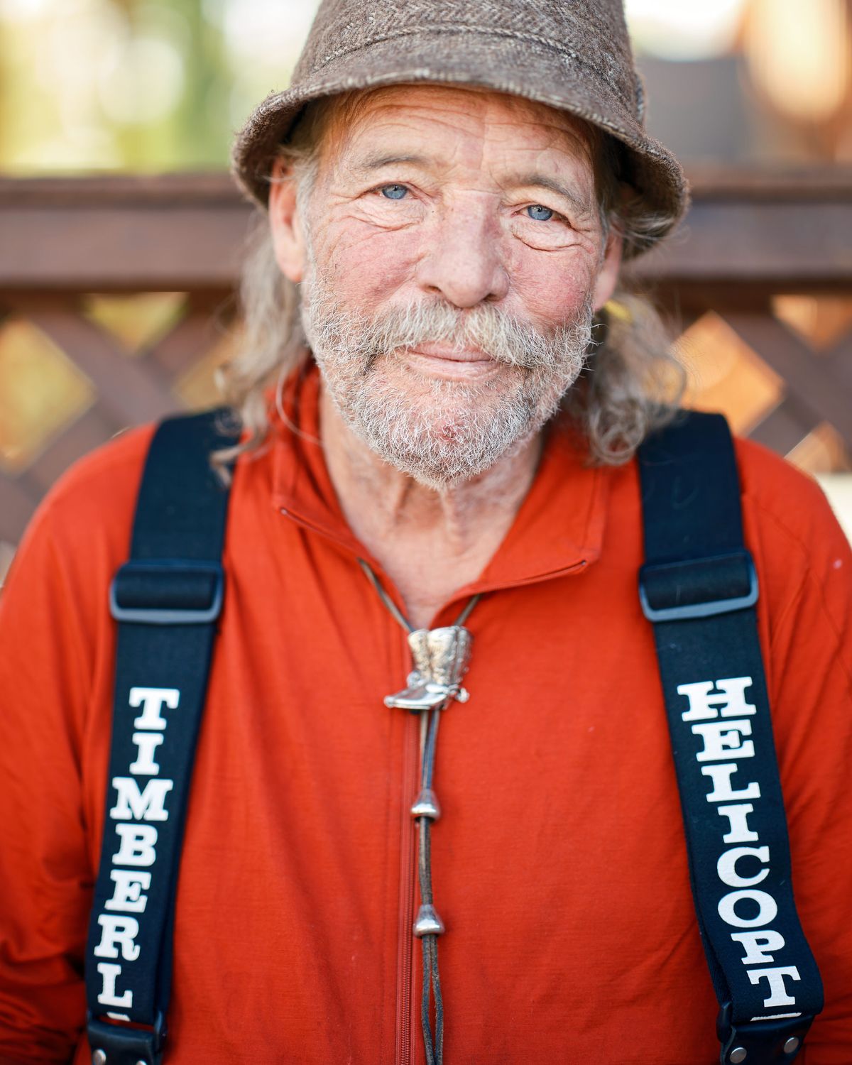 A portrait of forester and poet Mark Miller in Spray, Oregon. He is an older man with blue eyes, a grey beard, and long grey hair, wearing a red zip-up fleece, a grey bucket hat, and a silver cowboy boot bolo tie. Black suspenders printed with the words "TIMBERLINE" and "HELICOPTERS" cross his chest. He smiles warmly at the camera, with a wooden lattice fence and soft bokeh foliage visible in the background.