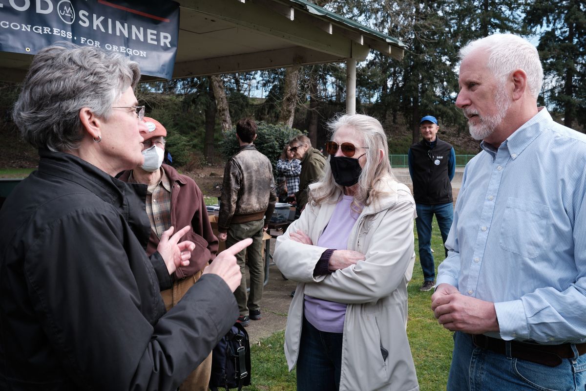 A group of people gathered outdoors at a political campaign event in Oregon City, Oregon, with a campaign banner visible in the background. The scene shows several individuals engaged in conversation, some wearing masks, in what appears to be a park setting with trees and grass.