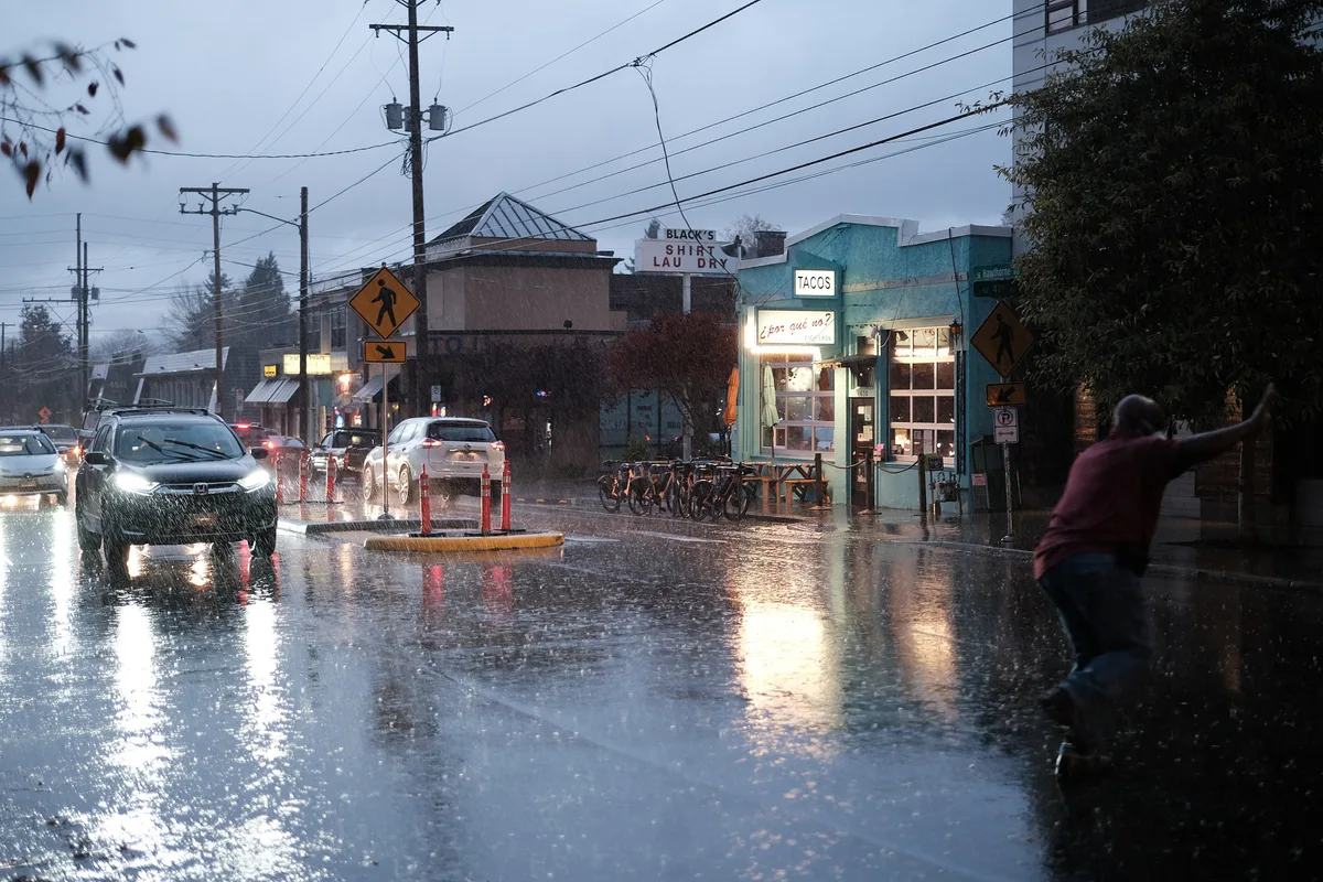 A heavy rainstorm drenches the intersection of SE Hawthorne Boulevard and SE 47th Avenue in Portland, Oregon. The teal-painted exterior of the popular ¿Por Qué No? taqueria glows warmly on the right, with its "Tacos" sign illuminated and a row of bikeshare bicycles parked out front. A man in a red shirt dashes across the flooded intersection on the right, arms outstretched for balance. Car headlights reflect off the standing water on the street as traffic backs up in the stormy blue-grey dusk. Black's Shirt Laundry is visible in the background.