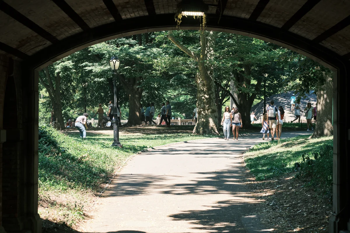 Framed by the shadowed arch of a stone bridge, families and couples emerge into dappled sunlight along Central Park's tree-lined pathways. The composition creates a natural vignette, where the dark tunnel opening contrasts dramatically with the bright summer scene beyond. Pedestrians stroll leisurely beneath towering canopies of green foliage, while children and adults gather on the sun-spotted lawns in this quintessential New York oasis. The interplay of light and shadow transforms an ordinary moment into a cinematic glimpse of urban park life.