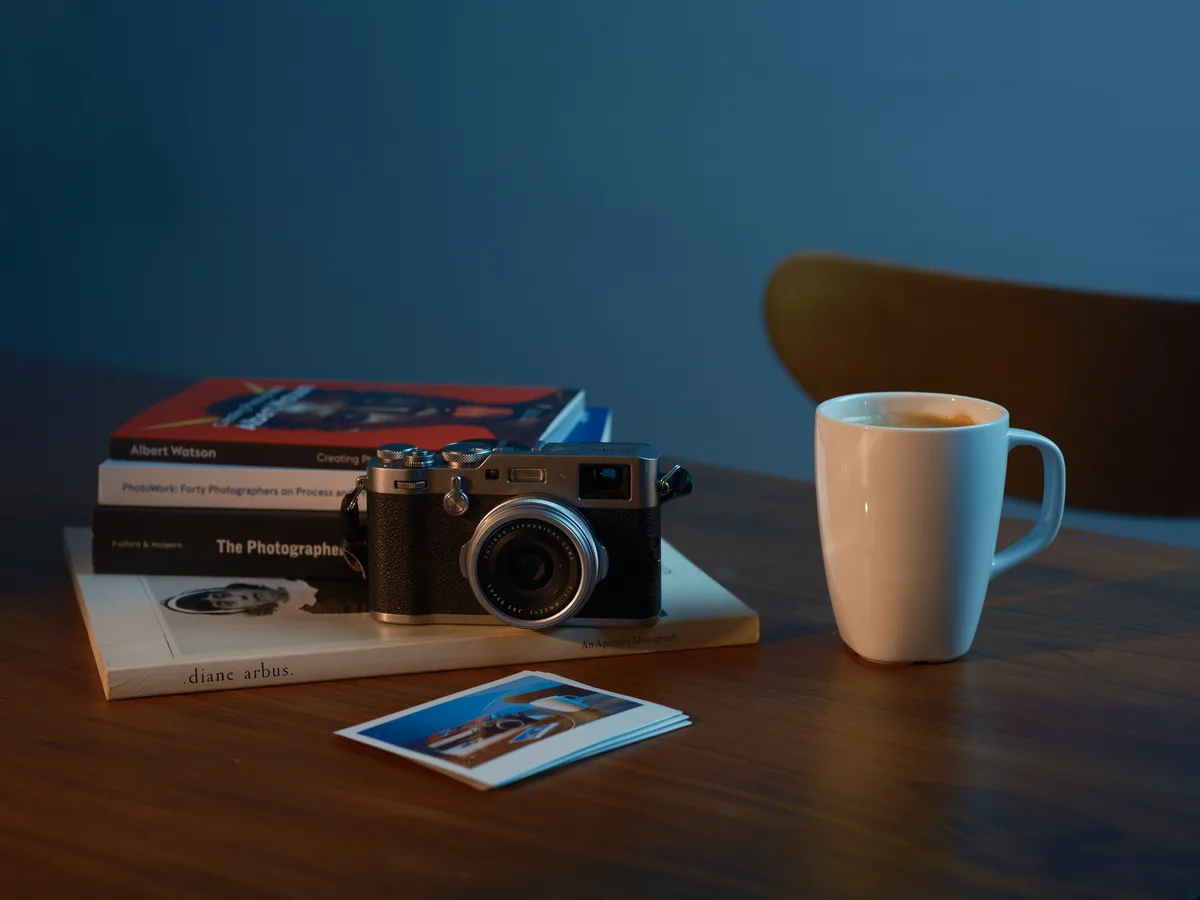 Golden hour light streams across a wooden desk in a Richmond District home, illuminating the tools of a contemplative photographer. A vintage rangefinder camera rests atop a carefully curated stack of photography books including works by Albert Watson and Diane Arbus, while a white ceramic mug holds steaming coffee nearby. Scattered color photographs lie casually beside the stack, suggesting a morning ritual of inspiration and caffeine in this Portland creative space.