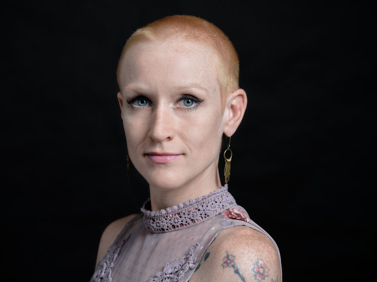 Against the deep charcoal backdrop of a Portland studio, Cayce Ryan commands attention with her striking platinum buzz cut and piercing blue eyes. Her delicate floral top, adorned with intricate lace details at the collar, creates a compelling contrast with her bold personal style and visible tattoo work. The professional headshot captures both vulnerability and strength, with soft studio lighting that emphasizes her direct gaze and the subtle pink tones of her lip color.