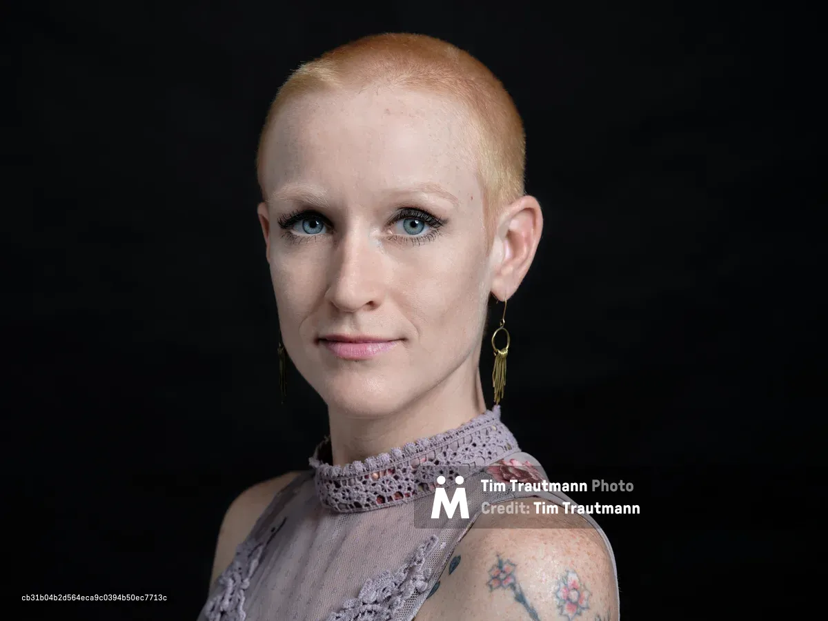 Against the deep charcoal backdrop of a Portland studio, Cayce Ryan commands attention with her striking platinum buzz cut and piercing blue eyes. Her delicate floral top, adorned with intricate lace details at the collar, creates a compelling contrast with her bold personal style and visible tattoo work. The professional headshot captures both vulnerability and strength, with soft studio lighting that emphasizes her direct gaze and the subtle pink tones of her lip color.