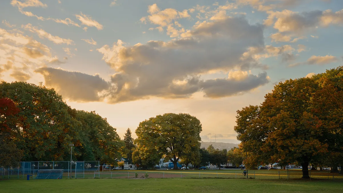 A sweeping autumn landscape at Powell Park in Portland, Oregon, bathed in warm golden evening light. Large mature trees in shades of green, amber, and rust frame an open grassy field with a baseball diamond and chain-link backstop visible on the left. A lone figure walks along the park's edge in the middle distance. Residential streets and parked vehicles are visible through the treeline, with the West Hills of Portland rising softly in the background. A dramatic sky of blue and gold-tinged clouds fills the upper half of the frame.