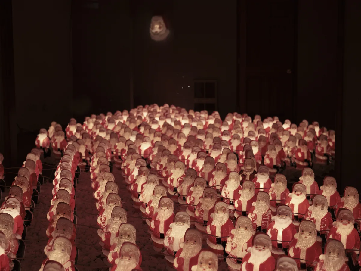 Hundreds of identical chocolate Santa figurines create a mesmerizing sea of red and white in Chris Willis's annual Portland art installation. The miniature holiday figures stretch across the darkened gallery floor in perfect rows, their faces catching warm artificial light that casts gentle shadows throughout the space. Above the sculptural army, a single moon-like orb glows mysteriously in the black void, creating an otherworldly atmosphere that transforms familiar Christmas imagery into something both whimsical and haunting.