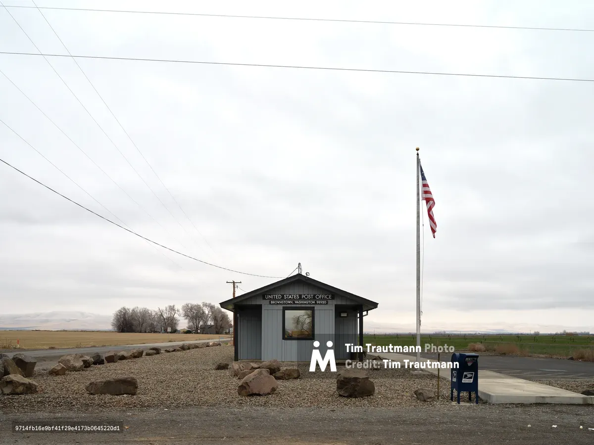 A weathered gray post office building stands solitary against the expansive Washington prairie, its patriotic signage declaring service to Brownstown's rural community. Overhead power lines slice through an overcast sky that mirrors the muted palette of basalt boulders scattered across the gravel foreground. The scene captures the essence of remote American infrastructure, where a simple structure and unfurled flag maintain connection to the wider world across miles of farmland. The composition emphasizes isolation and endurance, with the horizontal expanse of agricultural fields stretching toward distant mountains.