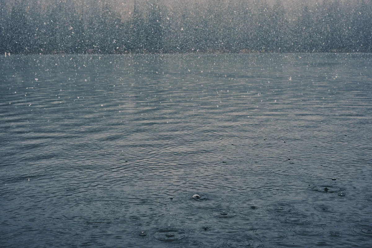 Countless raindrops create ephemeral ripples across the steel-gray surface of a Central Oregon lake near Sisters. The atmospheric precipitation transforms the water into a textured canvas of concentric circles, each drop's impact sending delicate rings outward before disappearing into the moody, overcast scene. The misty air above the lake blurs with falling rain, creating a dreamlike quality that speaks to the Pacific Northwest's contemplative weather patterns.