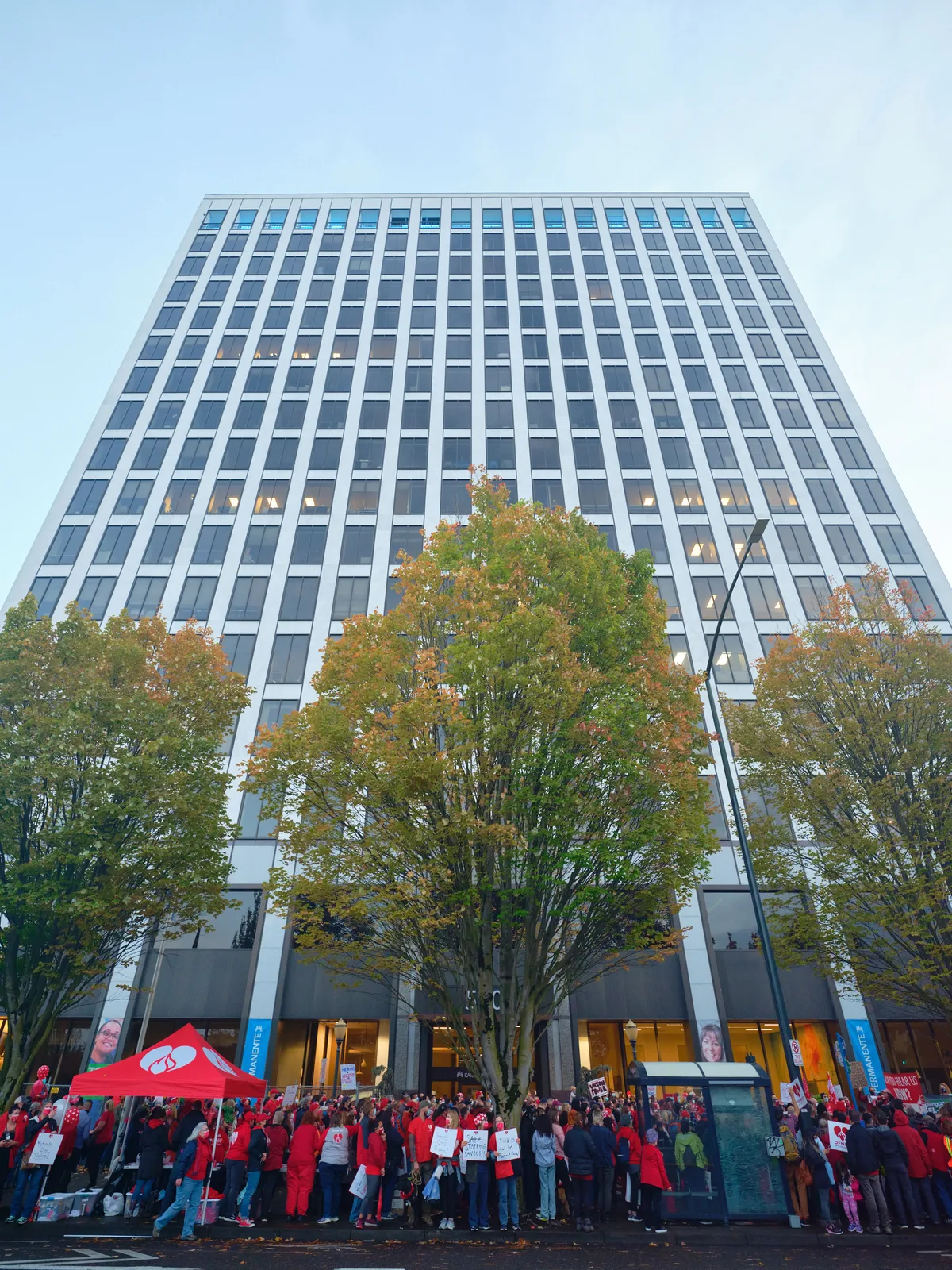 A sea of striking nurses in crimson scrubs and union red congregates at the base of Portland's imposing Kaiser Permanente Tower, their collective presence creating a powerful visual contrast against the building's stark modernist facade. The late afternoon light casts a cool blue wash over the scene while autumn trees frame the corporate monolith, their golden leaves echoing the warmth of the protesters' resolve. Signs and banners punctuate the crowd as healthcare workers mass in organized defiance, transforming the sterile corporate plaza into a stage for labor activism.