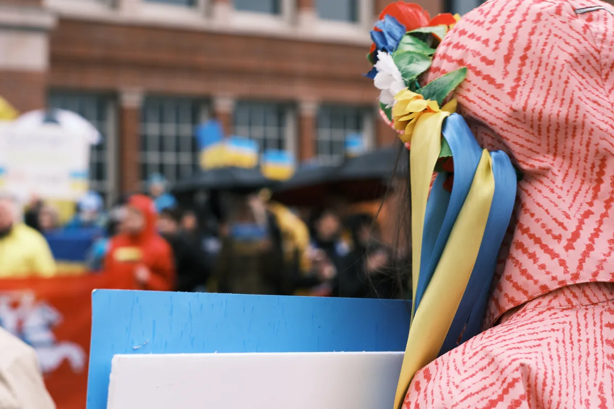 A woman in a coral-striped headscarf adorned with flowing blue and yellow ribbons—the colors of Ukraine—stands at a protest vigil outside Portland's Revolution Hall. The patriotic ribbons cascade from a crown of artificial flowers, creating a striking visual metaphor of hope and defiance. Behind her, blurred demonstrators gather in the urban streetscape, their forms dissolving into the shallow depth of field that isolates this singular moment of quiet determination.