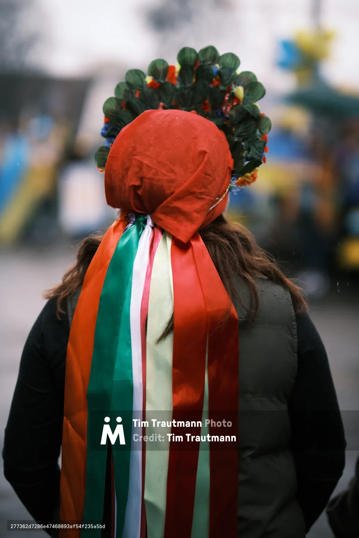 A solitary figure stands in defiant anonymity outside Portland's Revolution Hall, their identity shrouded by a crimson headwrap crowned with verdant leaves and beaded ornaments. Cascading ribbons in the colors of multiple flags—emerald, ivory, coral, and crimson—flow like liquid hope down their dark coat, creating a powerful symbol of international solidarity. The shallow depth of field dissolves the urban backdrop into impressionistic washes of blue and yellow, focusing attention on this poignant moment of pre-war protest in Southeast Portland's cultural district.