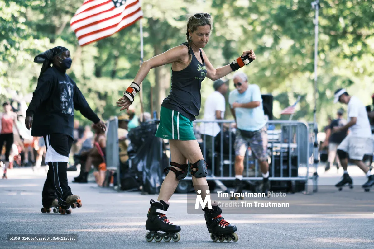 A determined female athlete glides across the asphalt of Central Park, her protective gear gleaming in the dappled afternoon sunlight. She moves with focused precision on inline skates, knee pads securing her stance as the American flag waves proudly in the soft-focused background. Spectators gather behind metal barriers, creating an atmosphere of community sport and urban recreation beneath the park's verdant canopy.