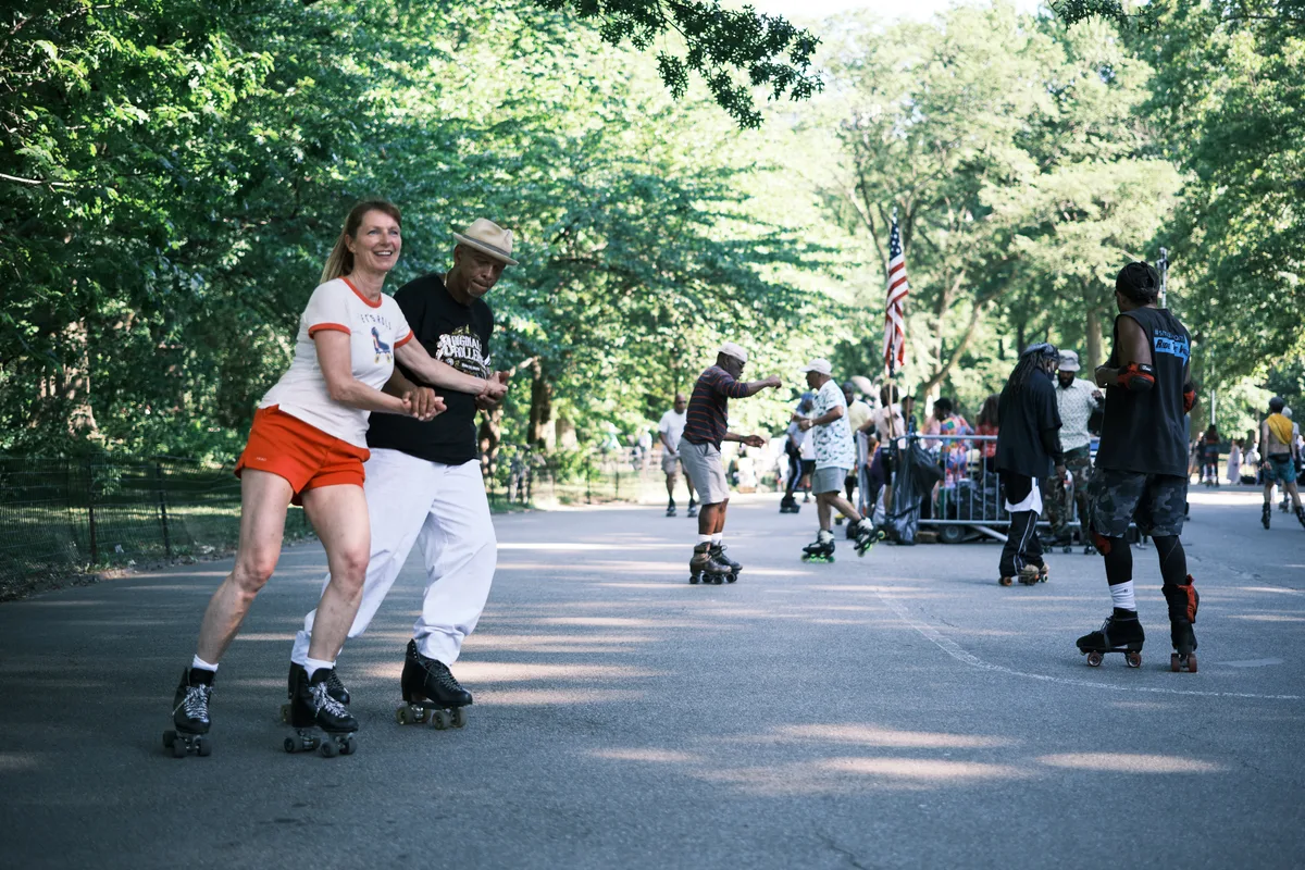 Two friends share a moment of pure delight as they roller skate together down a tree-lined pathway in Central Park. The woman in orange shorts and white shirt beams with laughter while holding hands with her companion in a beige hat and dark shirt, their synchronized movement captured against the dappled sunlight filtering through the lush summer canopy. Behind them, other skaters glide along the same path, creating a scene of community recreation beneath the park's towering trees. The warm, golden light and relaxed atmosphere perfectly capture the timeless appeal of roller skating culture in Manhattan's green heart.