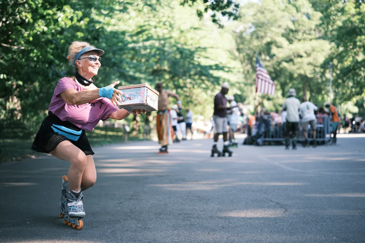 A spirited woman in a pink top and black athletic wear gracefully rollerskates along Central Park's tree-lined pathways, carrying what appears to be a lunch cooler. Dappled sunlight filters through the dense canopy above, casting dancing shadows on the asphalt as she navigates past blurred pedestrians and a distant American flag. Her confident posture and protective gear speak to both skill and safety consciousness in this quintessential New York moment.