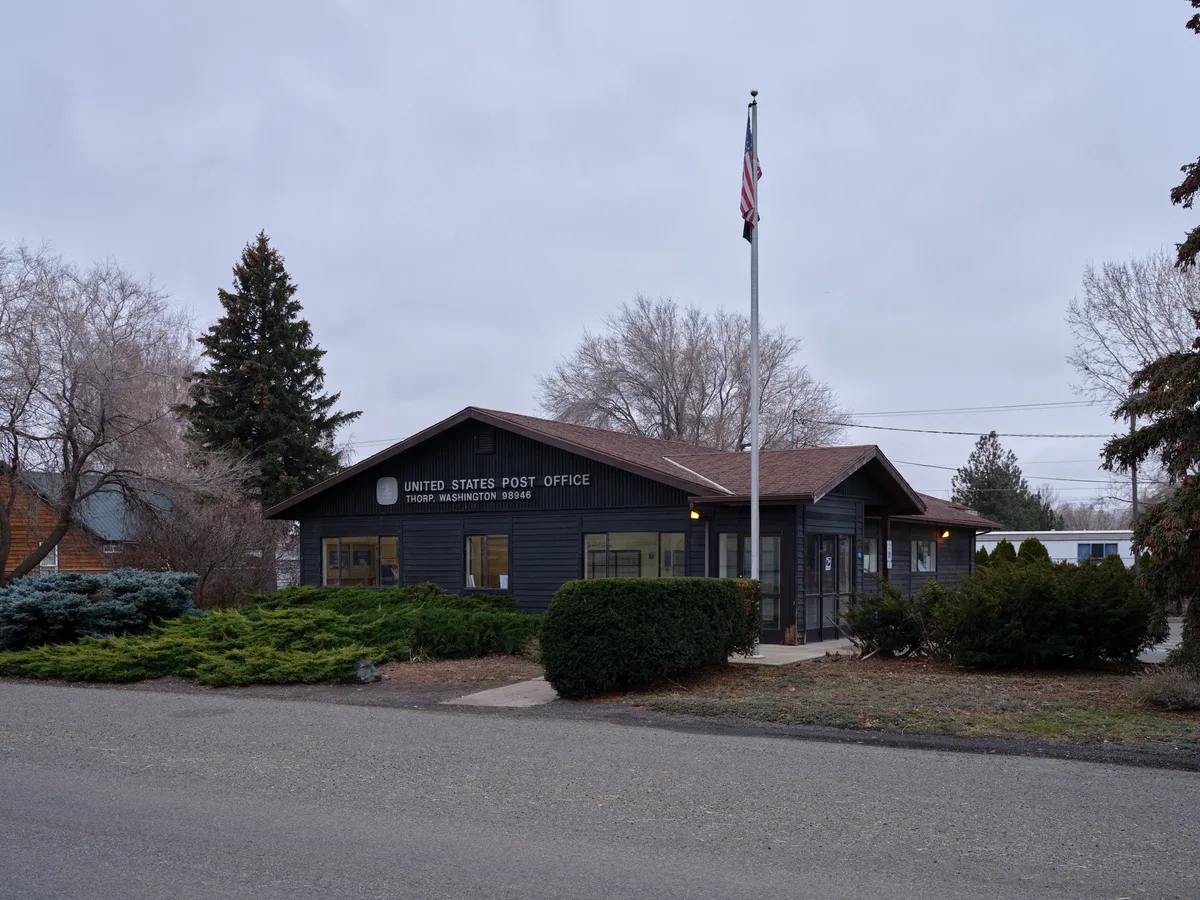 The modest United States Post Office in Thorp, Washington sits quietly beneath a pewter evening sky, its dark wood siding and warm interior glow creating an intimate portrait of small-town America. The single-story building, flanked by evergreen shrubs and bare winter trees, displays the Stars and Stripes on a tall flagpole that pierces the overcast horizon. Golden light spills from the windows onto the empty parking lot, suggesting the quiet continuity of postal service in this rural Kittitas County community.
