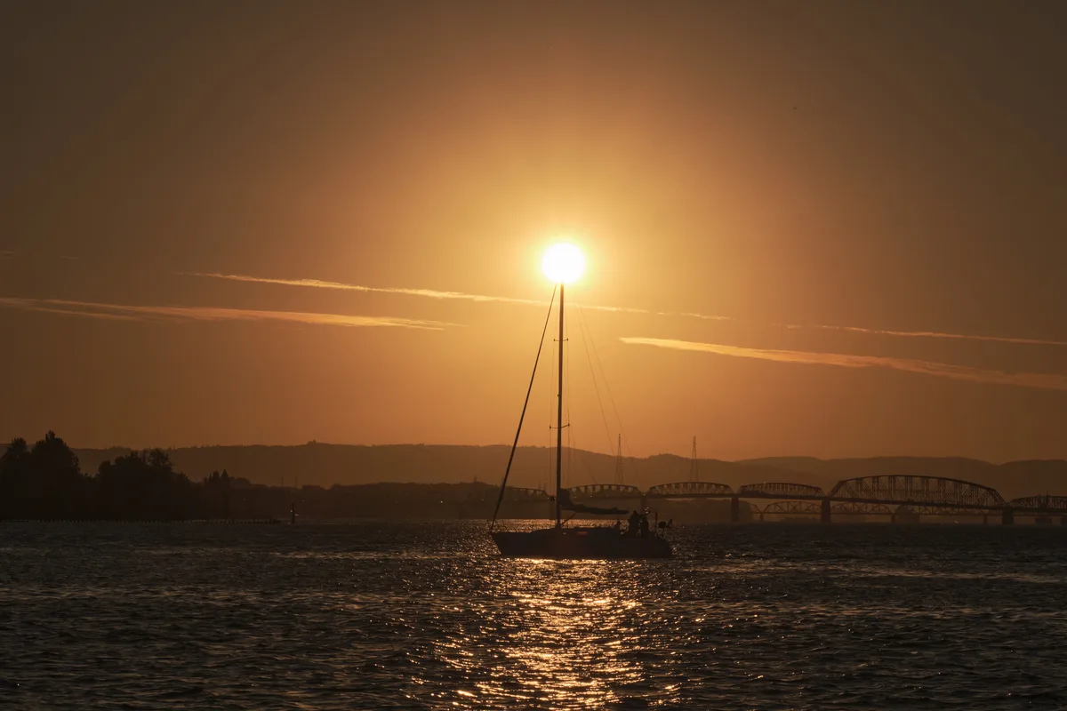 A silhouetted sailboat floats on calm water during golden hour sunset, with a large bridge visible in the background and the Columbia River reflecting warm sunlight in Portland, Oregon.