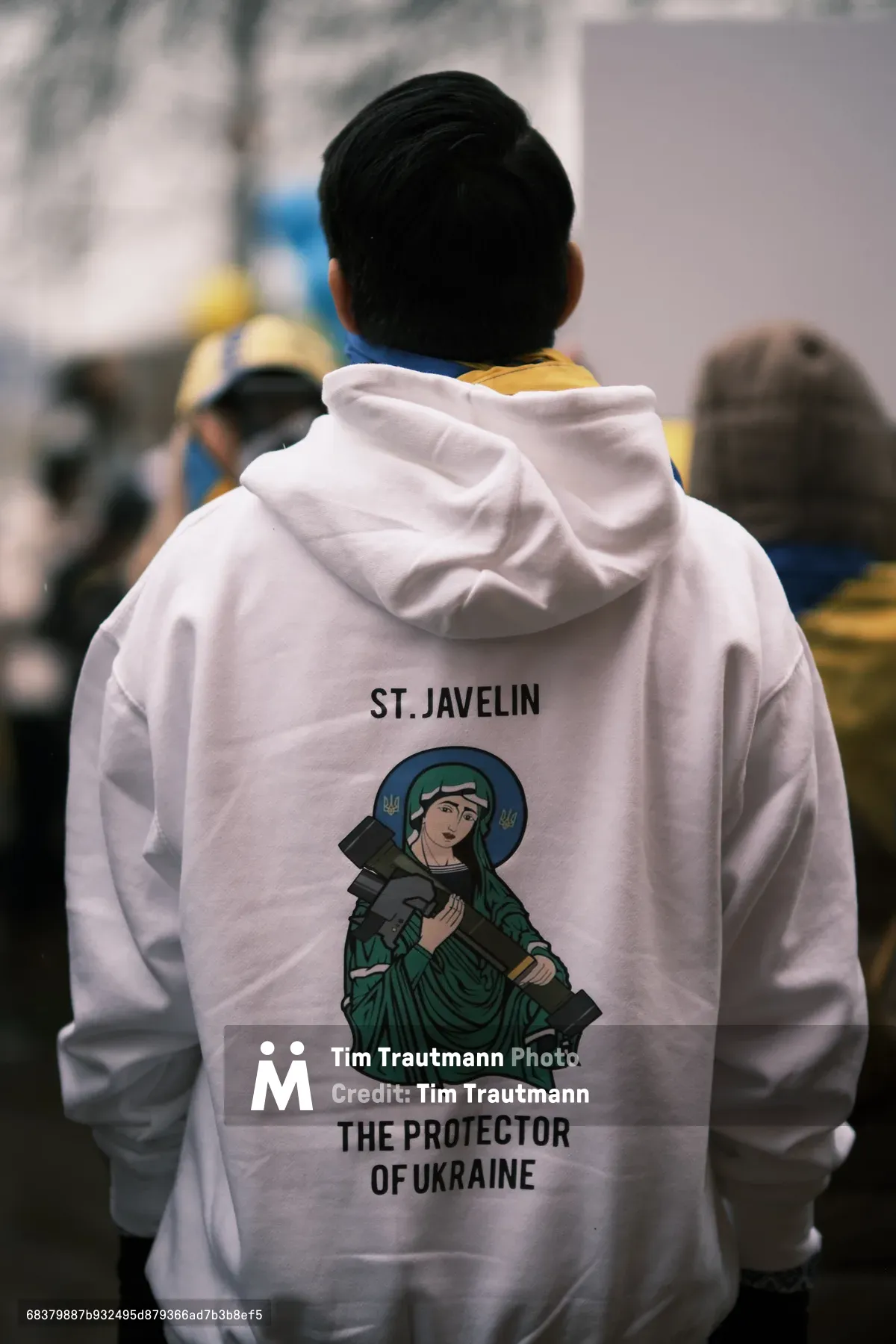 A figure in a white hoodie stands with their back to the camera outside Portland's Revolution Hall, displaying a striking illustration of St. Javelin—a folk saint depicted holding anti-tank weapons and labeled 'The Protector of Ukraine.' The shallow depth of field renders the surrounding crowd into a soft blur of muted colors, creating an atmosphere of quiet anticipation. The overcast Pacific Northwest light filters through bare winter branches, casting a somber mood over this prescient gathering that occurred just days before Russia's invasion of Ukraine began.