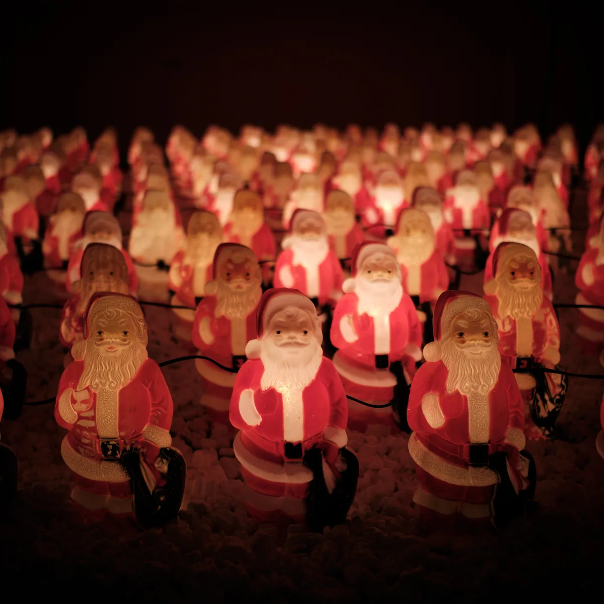 An enchanting nocturnal scene unfolds in Portland's Concordia neighborhood where hundreds of illuminated Santa Claus figurines create a surreal winter wonderland. The glowing plastic Santas, arranged in perfect rows, emanate a warm amber light that pierces the velvety darkness, their jolly faces creating an almost hypnotic pattern that stretches into the shadows. Chris Willis's annual Santa Clones installation transforms this Northeast Portland corner into a whimsical spectacle, where mass-produced holiday cheer takes on an unexpectedly artistic quality. The soft bokeh effect in the background dissolves the distant figures into dreamlike orbs of red and gold, creating depth and mystery in this delightfully surreal Christmas tableau.