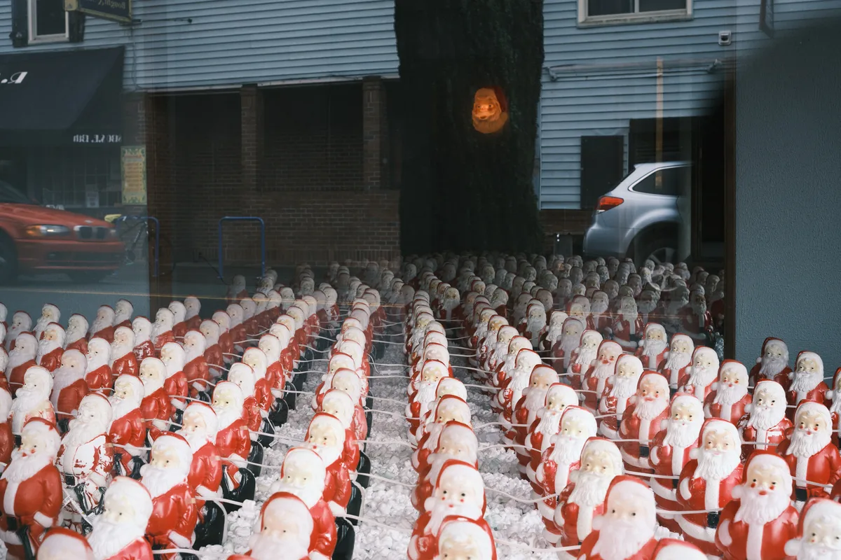 Hundreds of identical Santa Claus figures occupy the floor of Consignment Store in Portland's Sellwood neighborhood, creating Chris Willis's surreal annual art installation. The ceramic or plastic Santas are arranged in precise rows on wire shelving, their jolly faces creating an unsettling sea of mass-produced Christmas cheer. Gallery windows reflect the quiet street outside, where a silver SUV and storefronts hint at the ordinary world beyond this dreamlike tableau of holiday multiplication.