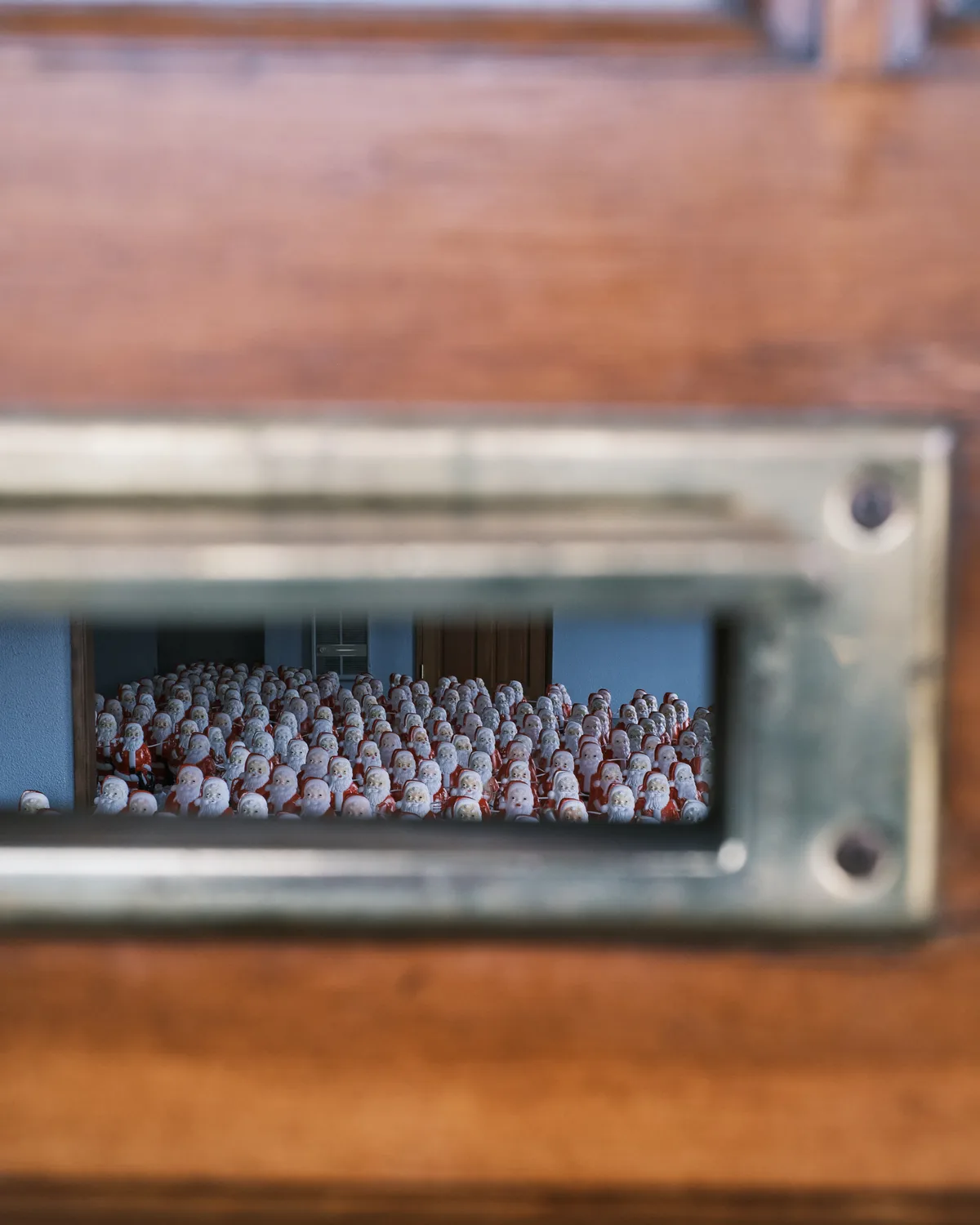 Through the narrow aperture of a gallery mailslot, hundreds of identical Santa figurines crowd together in ghostly formation, their red-suited forms creating a sea of holiday repetition. The voyeuristic perspective frames this uncanny army of Christmas icons within the confines of Portland's Consignment Gallery, where artist Chris Willis's annual installation transforms the familiar into the surreal. Warm wood tones of the gallery exterior contrast sharply with the cool interior lighting that illuminates this manufactured multitude, creating an intimate yet unsettling glimpse into mass-produced festivity.