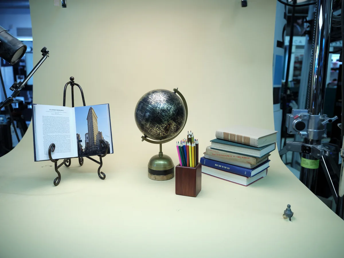 A meticulously arranged still life captures the essence of intellectual pursuit on a pristine white surface within a professional photography studio. The composition centers around a dark celestial globe with golden meridian rings, flanked by a stack of weathered books, an open architectural volume displaying the iconic Flatiron Building, and a wooden pencil holder bristling with colorful writing instruments. Studio lighting creates subtle shadows and highlights against the neutral backdrop, while professional equipment frames the edges, revealing the careful orchestration behind this contemplative tableau.