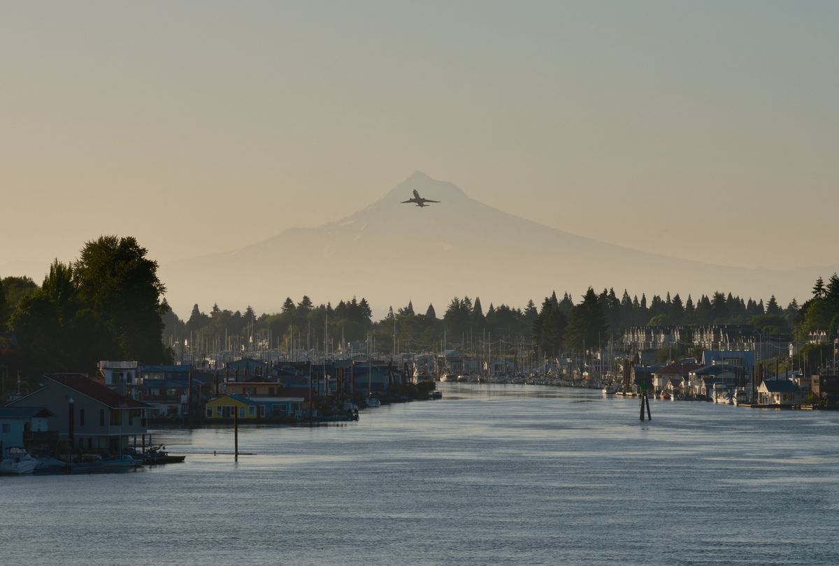 A airplane flies directly over a snow-capped mountain peak in the distance, with a marina full of boats and houseboats in the foreground under a hazy, golden-hour sky. The scenic waterway is lined with evergreen trees and various watercraft creating a peaceful Pacific Northwest landscape.