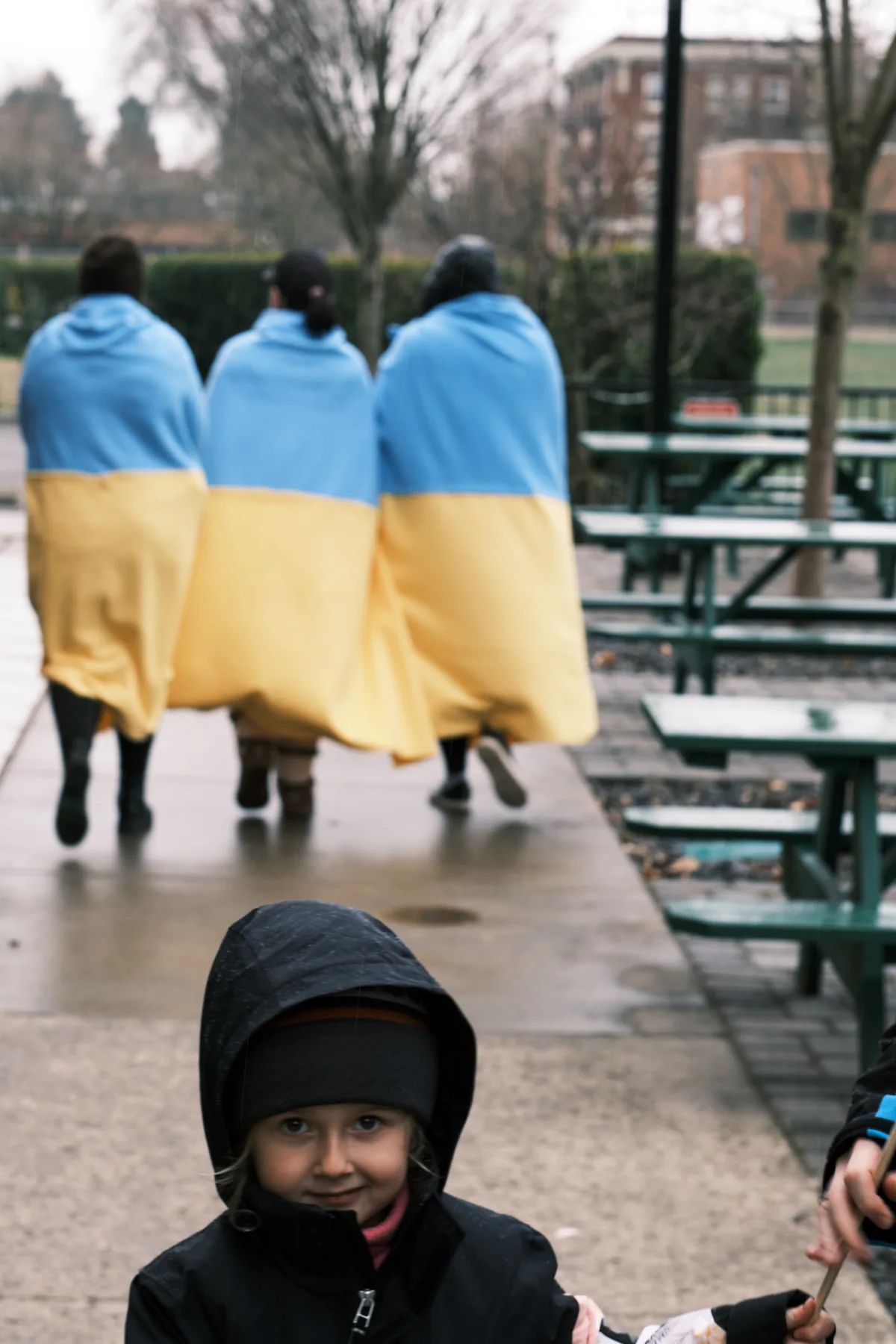 Three figures draped in Ukrainian flags stand with their backs turned on the rain-slicked plaza outside Portland's Revolution Hall, their blue and yellow shrouds creating a haunting tableau of solidarity. A young child in a dark hooded jacket sits in the foreground, gazing directly at the camera with knowing eyes that belie their years. The overcast February sky and bare winter trees frame this prescient moment of protest, captured in the days before Russian forces would cross into Ukraine.