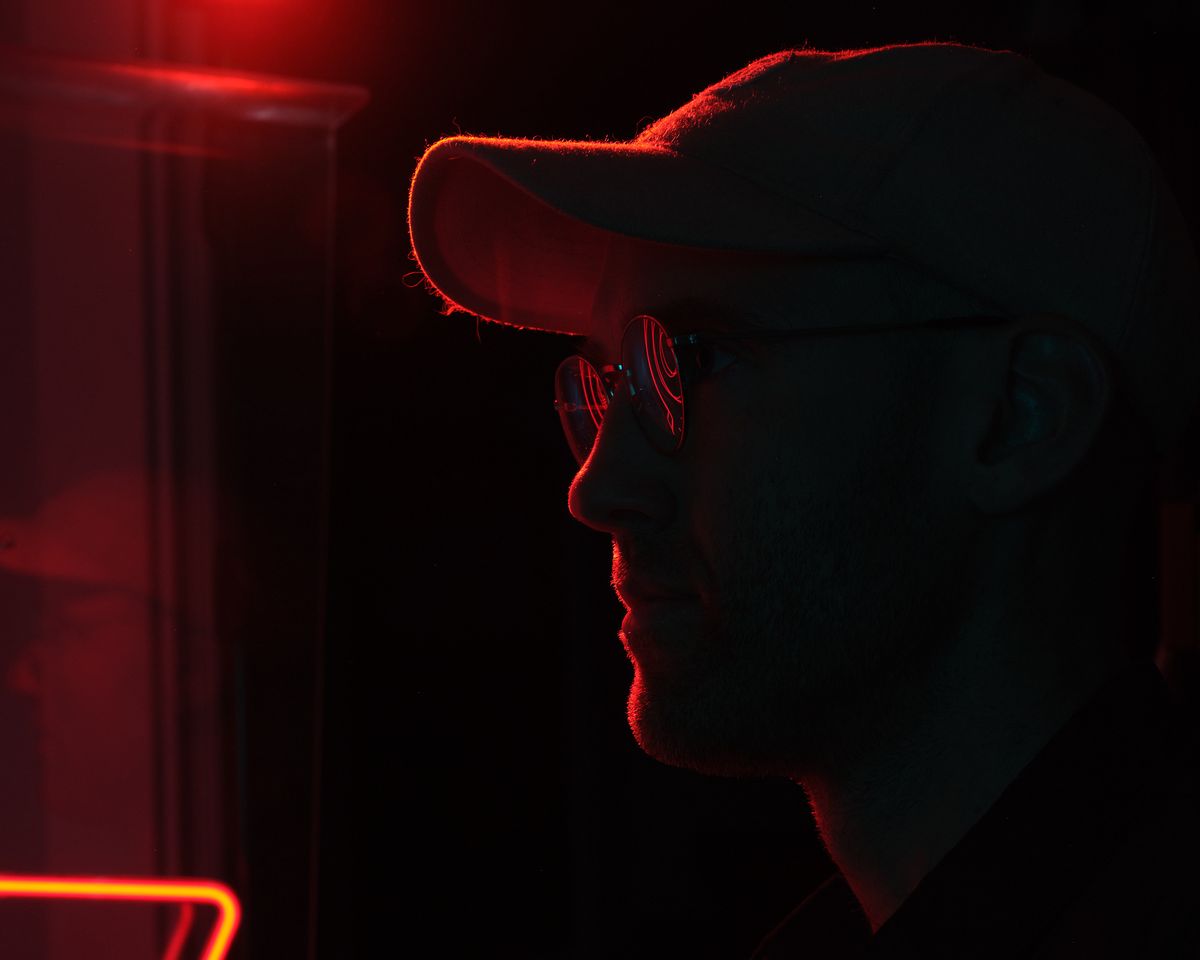 A dramatic silhouette of a person wearing a hat and sunglasses, illuminated by intense red neon lighting against a dark background. The moody lighting creates strong contrast and rim lighting effects around the subject's profile.