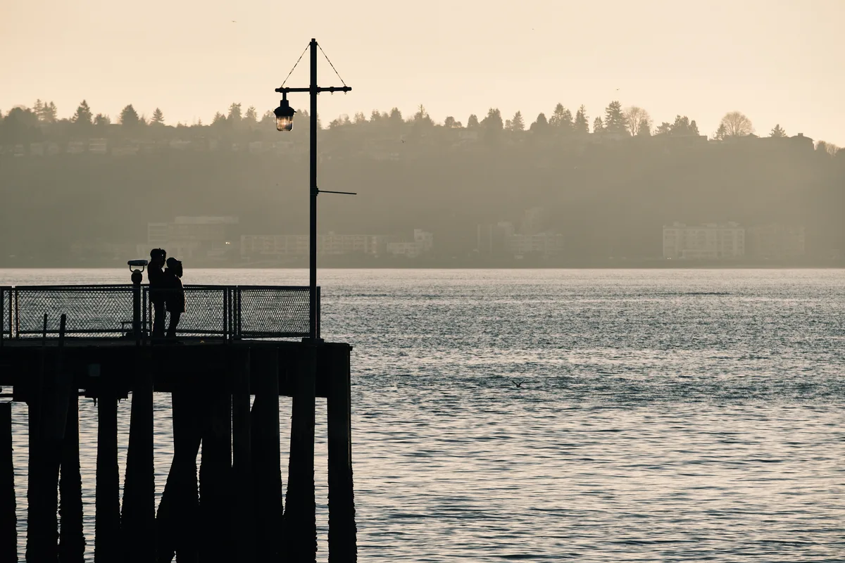 Two figures stand in intimate silhouette against the amber-washed waters of Elliott Bay from Seattle's Pier 70, their forms darkened against the ethereal golden hour light. A vintage lamp post rises beside them like a sentinel, while the distant shoreline dissolves into layers of atmospheric haze. The weathered wooden pier pilings create strong vertical lines that anchor the composition, as gentle ripples catch the dying light across the bay's surface.