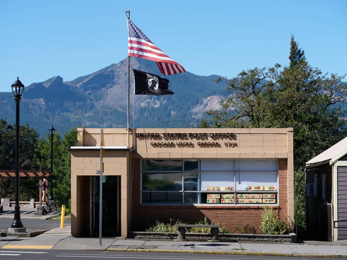The mid-century brick and concrete United States Post Office stands sentinel in Cascade Locks, Oregon, its modernist lines softened by the dramatic backdrop of forested mountains rising into crystal blue sky. Two flags—the Stars and Stripes and a black POW/MIA banner—flutter from a single pole, while vintage street lamps frame the humble federal building. The Columbia River Gorge's towering peaks create a theatrical backdrop for this essential piece of small-town American infrastructure, where community connection meets governmental service.