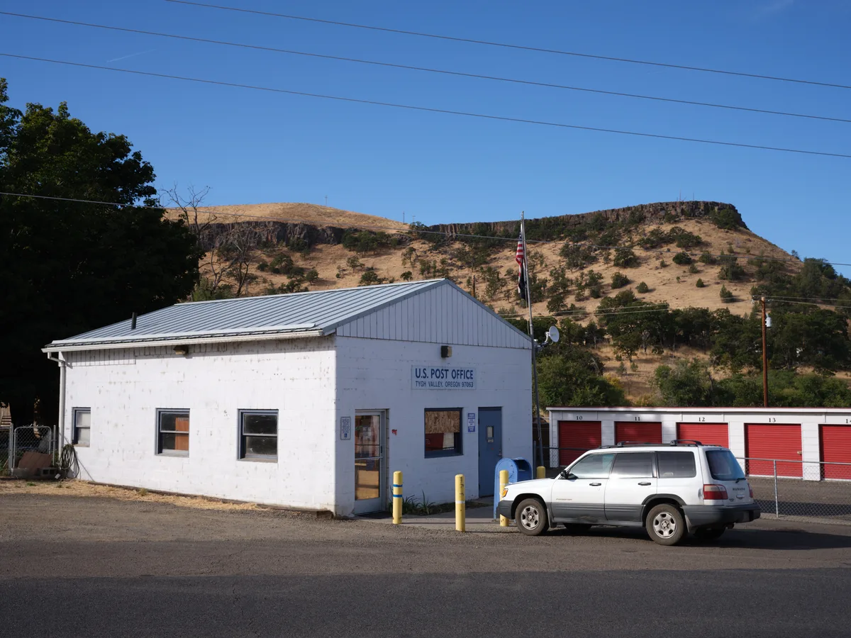 A modest white-painted U.S. Post Office building sits beneath the dramatic golden bluffs of Tygh Valley, Oregon, its metal roof gleaming under the crystalline blue sky. The humble single-story structure, flanked by red storage units and a weathered SUV, embodies the essential services that anchor rural American communities. Power lines streak across the azure expanse above sun-baked hills dotted with sparse vegetation, while shadows from mature trees provide relief from the high desert light. The scene captures the quiet dignity of small-town infrastructure against the raw beauty of the Columbia River Gorge region.