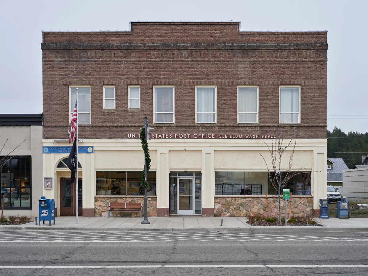 A weathered brick United States Post Office stands resolute along East 1st Street in Cle Elum, Washington, its classic early 20th-century facade crowned with traditional red brick and cream-colored stone trim. The overcast Pacific Northwest sky casts even light across the building's seven tall windows, while the American flag hangs quietly beside the entrance. Stone wainscoting and large storefront windows reveal the building's dual role as both federal facility and community anchor, set against the backdrop of the Cascade Mountains' forested hills.