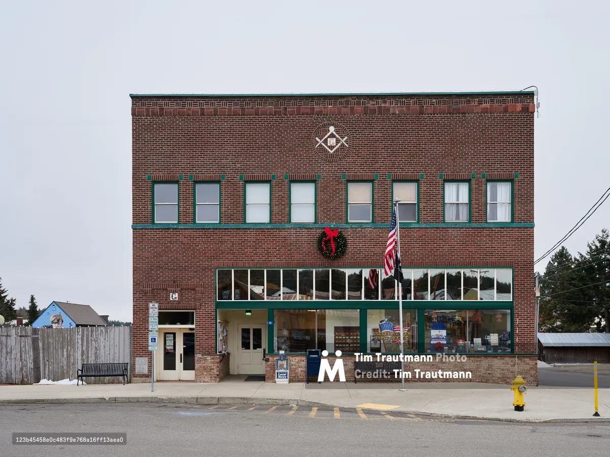 A classic two-story brick building houses the United States Post Office in Roslyn, Washington, its weathered red facade adorned with green window trim and a festive holiday wreath. The Masonic lodge emblem crowns the upper story while an American flag stands sentinel beside the entrance. Under an overcast winter sky, this quintessential small-town structure embodies the enduring presence of federal service in rural America, its large storefront windows revealing the community lifeline within.