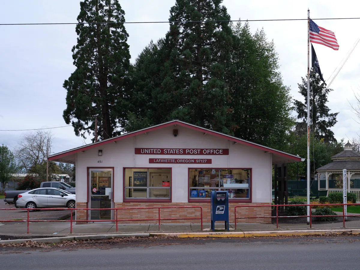 A modest single-story post office building stands beneath towering evergreens in the small Oregon town of Lafayette, its cream-colored walls accented with burgundy trim and signage. The American flag waves proudly from a tall pole beside the building, while power lines stretch across an overcast sky that speaks to the Pacific Northwest's characteristic atmosphere. A blue USPS mailbox sits sentinel on the sidewalk, and cars parked nearby suggest the steady rhythm of daily community life in this rural Yamhill County settlement.