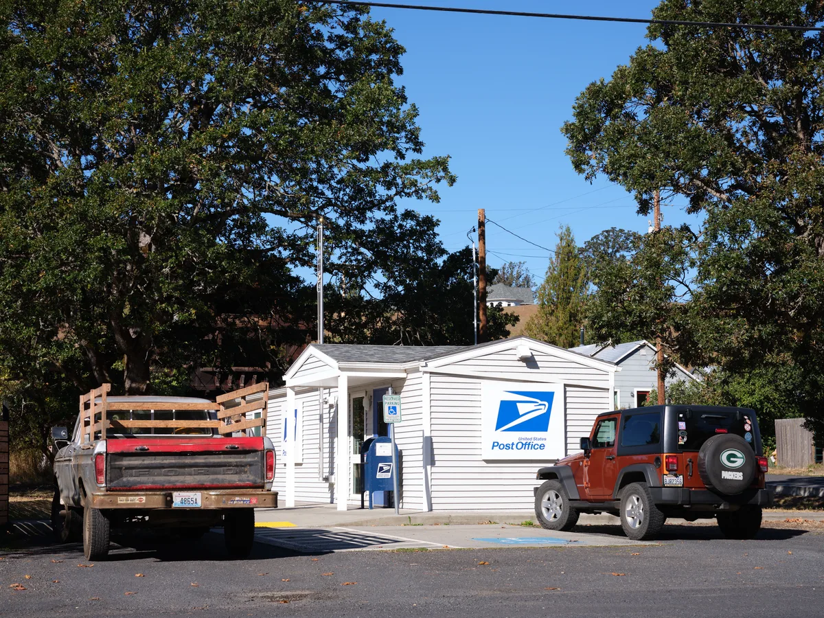 A modest white clapboard post office building sits nestled among towering mature trees in Husum, Washington, embodying the essence of small-town America. The single-story structure displays the iconic blue USPS logo prominently on its facade, while a blue mailbox stands sentinel beside the entrance. Two vehicles - a weathered pickup truck and an orange Jeep - are parked in front, suggesting the steady rhythm of daily community life under a brilliant azure sky.