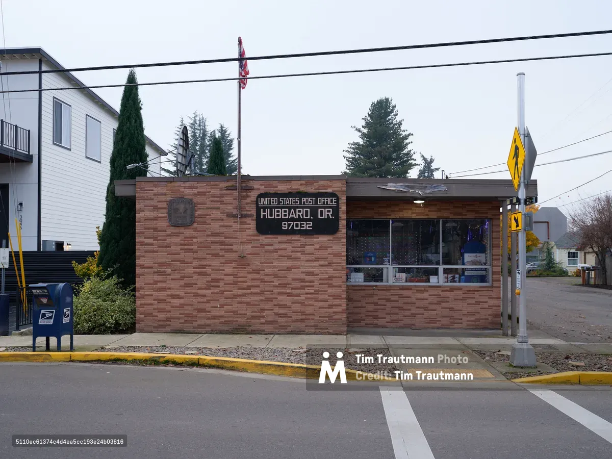 The modest brick facade of the United States Post Office in Hubbard, Oregon stands resolute against a pewter sky, its weathered red-brown masonry speaking to decades of faithful service. Power lines stretch across the frame like musical staves, while the yellow diamond traffic sign and blue mailbox punctuate the muted palette with primary colors. The institutional architecture blends seamlessly into this quiet Marion County streetscape, where modern siding and mature evergreens frame this essential civic cornerstone.