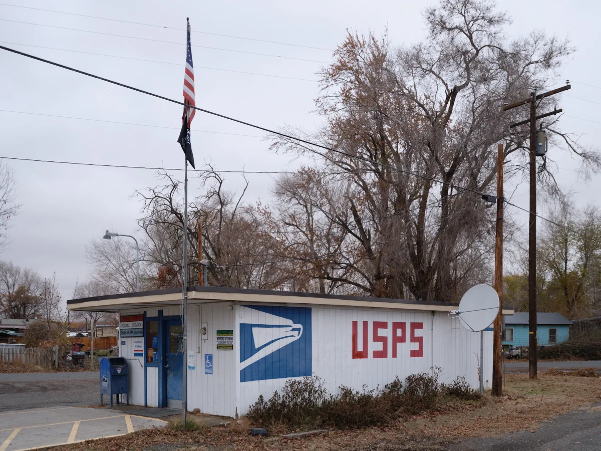 A modest USPS facility stands beneath overcast winter skies in Parker, Washington, its white metal siding adorned with the familiar blue postal service logo and red lettering. Bare cottonwood trees loom behind the single-story building, their leafless branches creating a stark silhouette against the pewter sky. An American flag hangs motionless from a utility pole, while power lines crisscross the frame, emphasizing the rural isolation of this essential community service hub.