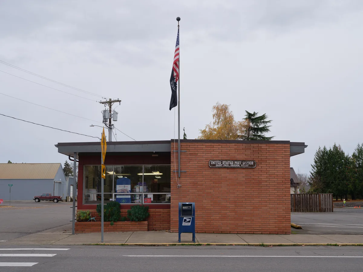 A modest brick United States Post Office stands sentinel in Saint Paul, Oregon, its mid-century modern architecture crowned by the American flag and POW-MIA banner fluttering against pewter-gray clouds. The building's warm red brick facade contrasts with the somber autumn sky, while a lone blue mailbox guards the sidewalk and utility lines sketch geometric patterns overhead. The scene captures the quiet dignity of rural American infrastructure, where community services endure beneath the Pacific Northwest's characteristic overcast atmosphere.