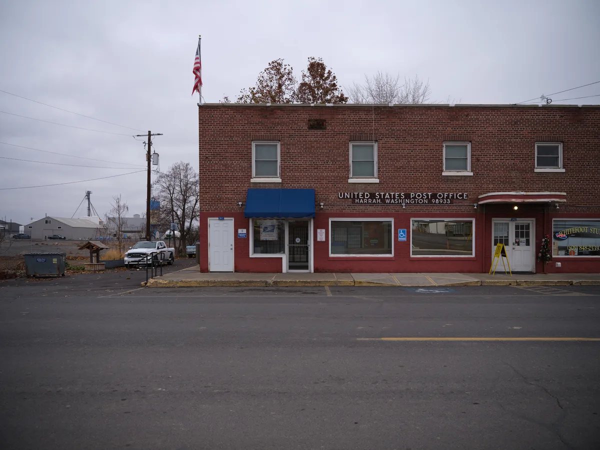 A modest brick United States Post Office building stands along a quiet street in Harrah, Washington, its red brick facade weathered by time and Pacific Northwest rains. The overcast sky casts a muted, contemplative light across the scene, while bare autumn trees frame the institutional building with its distinctive blue awning and official signage. Power lines stretch overhead, connecting this rural postal outpost to the broader community it serves.