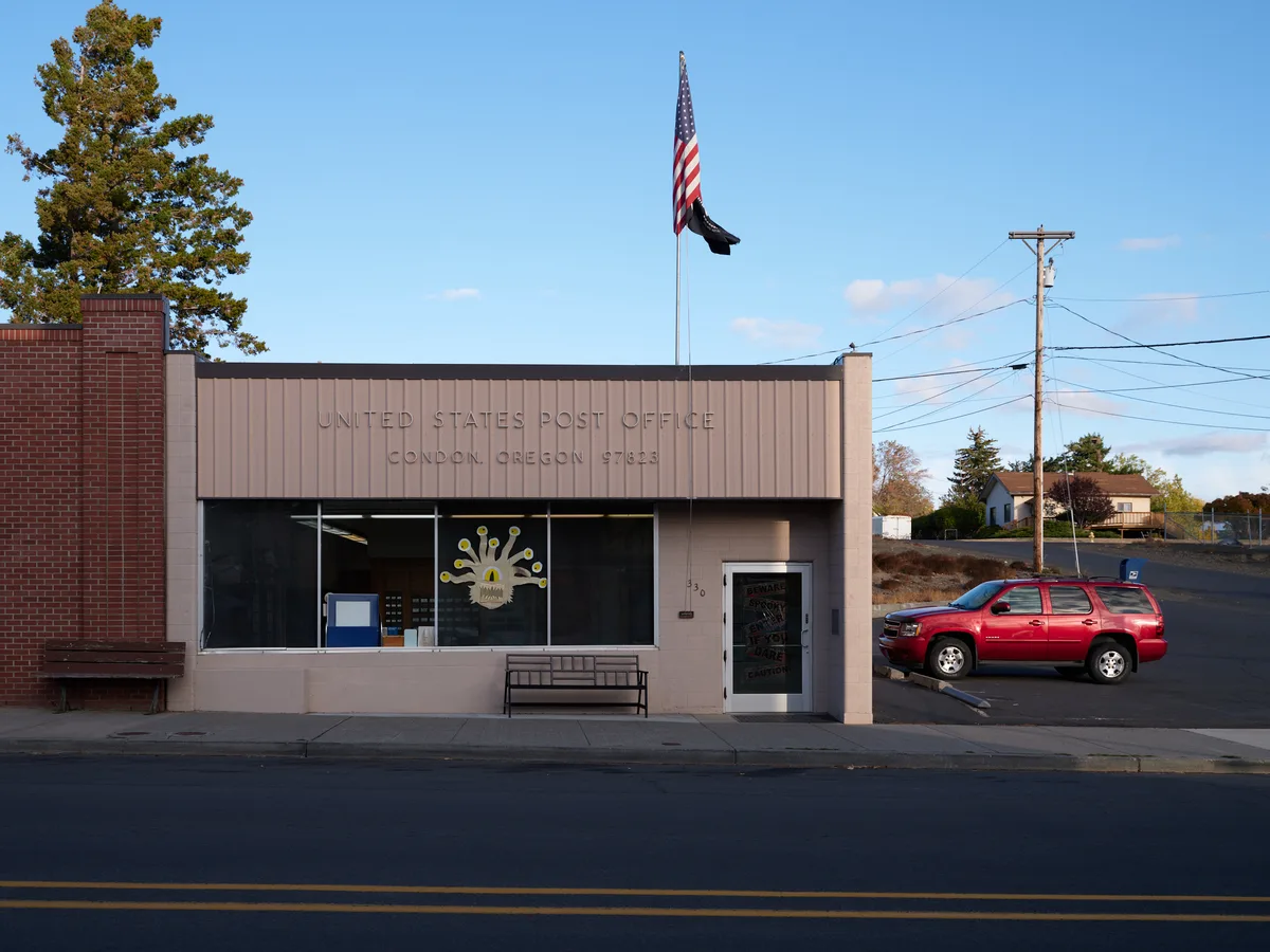 Golden hour light bathes the modest United States Post Office in Condon, Oregon, where mid-century institutional architecture meets small-town practicality. The beige corrugated metal facade displays the building's federal purpose in simple block letters, while a red Chevrolet SUV sits sentinel in the adjacent parking lot beneath a proud American flag. Power lines slice through the crystalline blue sky above this essential community hub, where autumn trees frame the scene with seasonal warmth in Oregon's rural Gilliam County.
