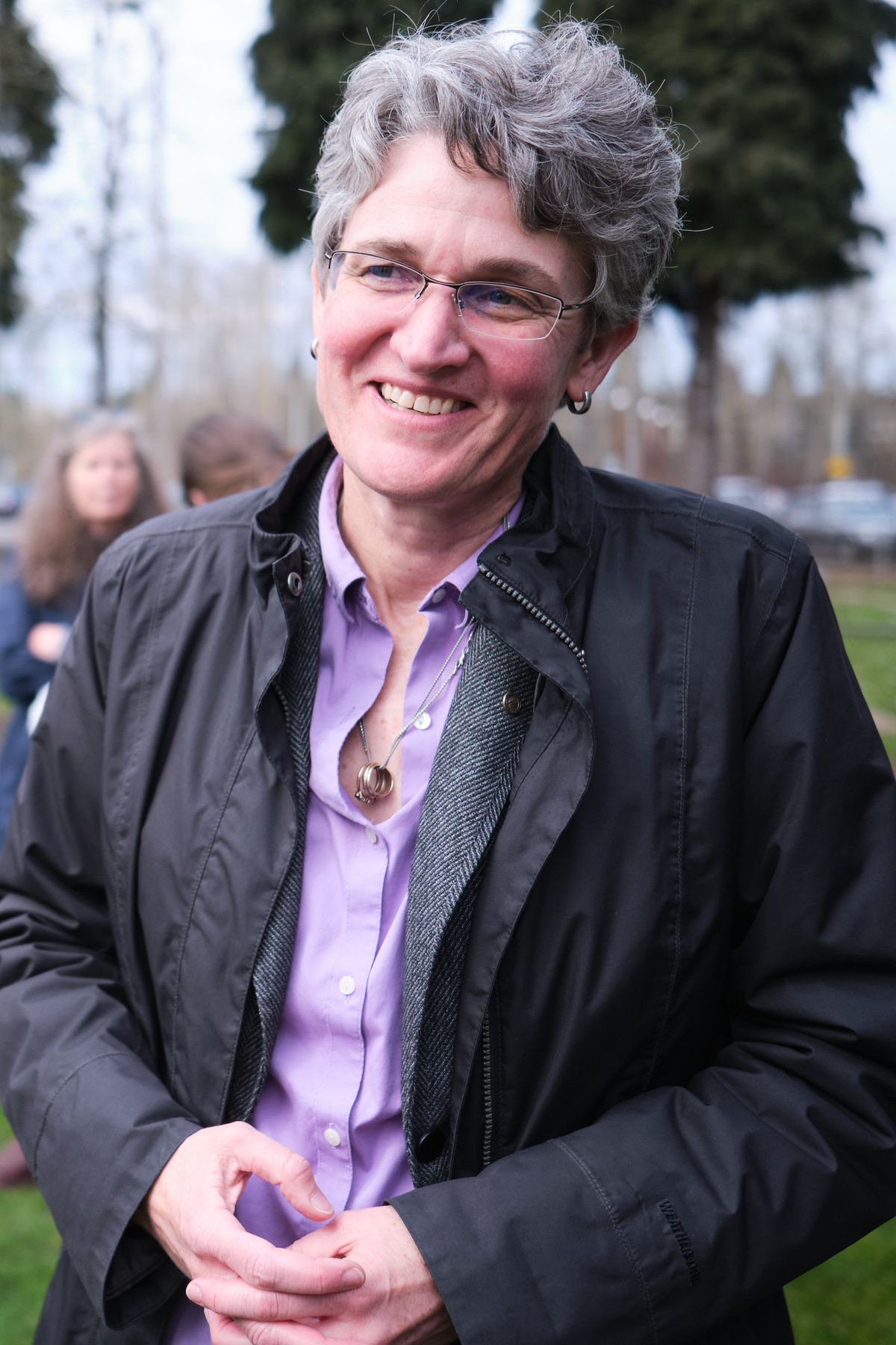 A middle-aged woman with curly gray hair and glasses smiles warmly at the camera while wearing a purple button-up shirt under a dark jacket. She has her hands clasped together and is photographed outdoors in what appears to be a park or community gathering space.