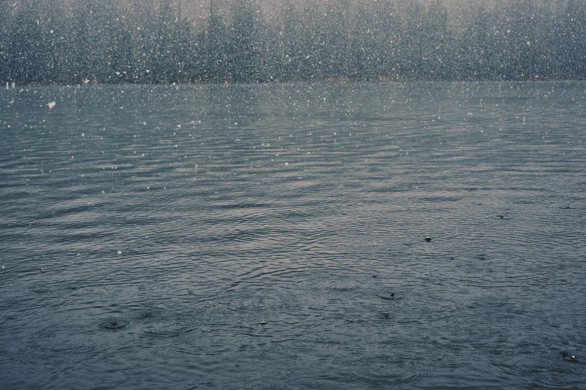 Heavy snowfall creates a curtain of white across a steel-gray lake surface in the Oregon Cascades near Sisters. The atmospheric precipitation transforms the water into a textured canvas of ripples and droplets, while the distant shoreline dissolves into a soft, monochromatic haze. The image captures the raw intensity of a mountain winter storm, with each snowflake visible as delicate points of light against the moody, overcast sky.