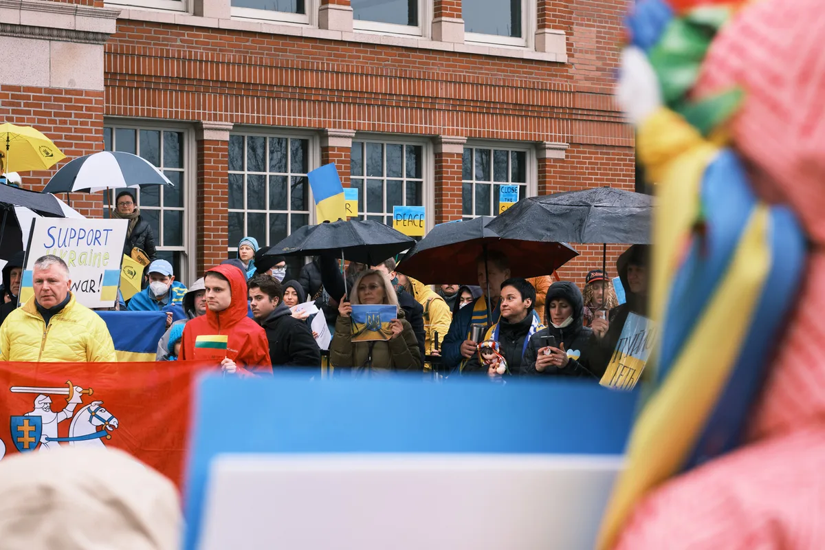 Rain-soaked demonstrators huddle beneath black umbrellas outside Portland's historic Revolution Hall, their blue and yellow Ukrainian flags creating vivid splashes of color against the red brick facade. Signs reading "SUPPORT" and "PEACE" pierce through the crowd as protesters gather in defiant solidarity days before Russian hostilities would begin. The overcast sky and wet pavement amplify the urgency of their message, while the stately Buckman neighborhood building provides a dignified backdrop to this prescient moment of civic engagement.