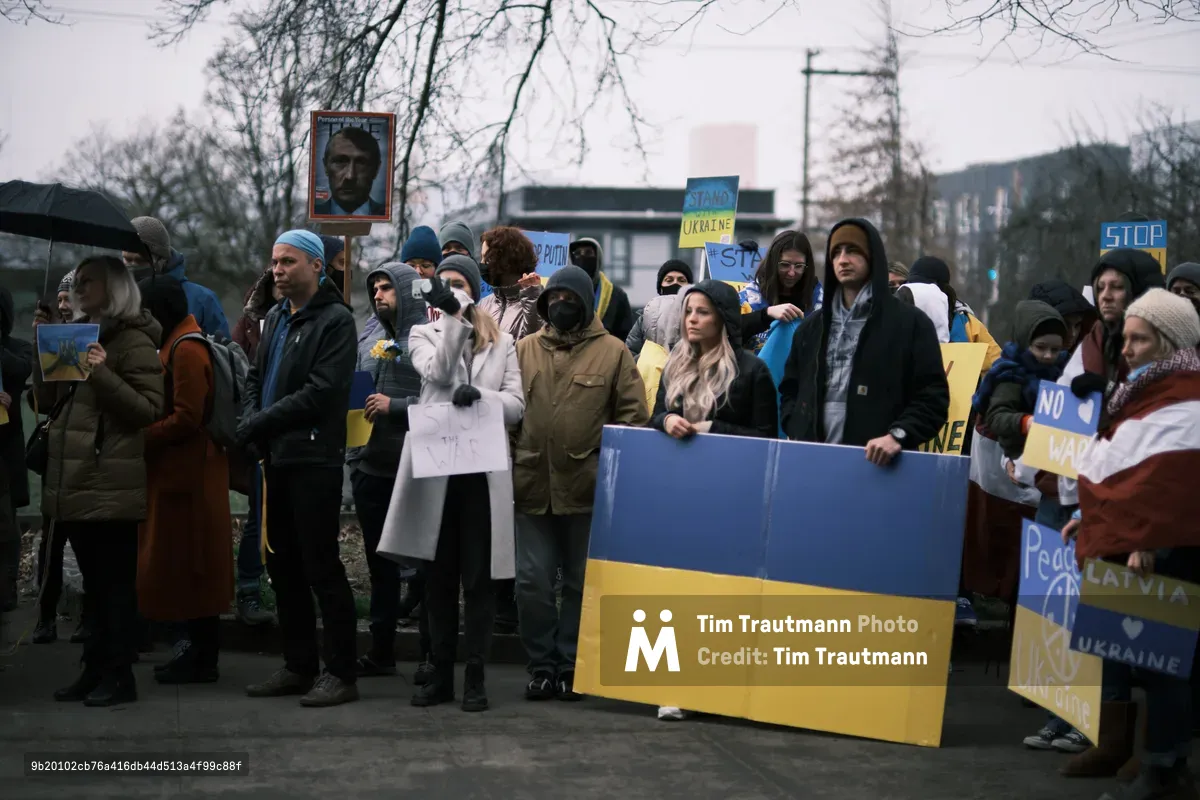 Bundled against Portland's winter chill, protesters congregate outside Revolution Hall on Southeast Stark Street, their blue and yellow Ukrainian flags creating stark splashes of color against the overcast February sky. The diverse crowd of activists holds handmade signs and portraits, their solemn faces reflecting the gravity of impending conflict as bare tree branches frame this moment of international solidarity. The muted daylight casts a prophetic atmosphere over this prescient demonstration, captured just days before Russia's invasion would transform their peaceful protest into tragic prophecy.