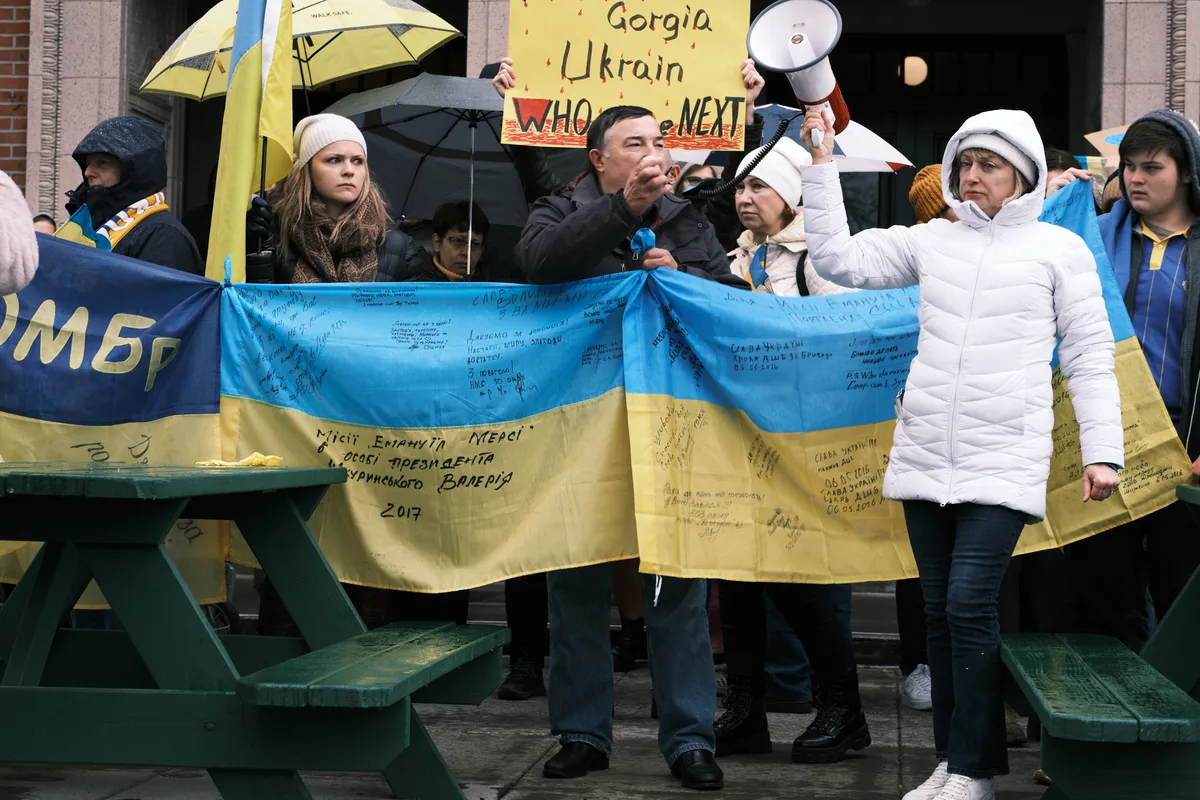 Ukrainian demonstrators gather on the weathered steps of Portland's Revolution Hall, their hands gripping a signature-laden blue and yellow flag that ripples with the weight of collective hope and dread. A man in dark clothing speaks passionately into a microphone while protesters in white winter coats stand resolute behind him, yellow umbrellas blooming overhead like defiant flowers against the overcast Pacific Northwest sky. Signs reading 'Georgia Ukraine WHO is NEXT' pierce through the crowd, their urgent handwriting capturing the prescient anxiety of a community bracing for war.