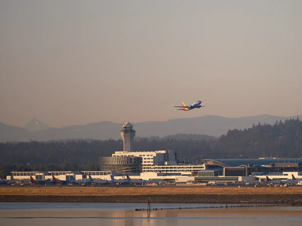 A Southwest Airlines aircraft takes off against a dramatic sunset sky above the Portland International Airport terminal and control tower, with forested hills and mountains visible in the background and calm water in the foreground.